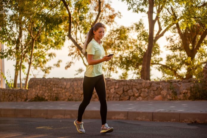 Close-up portrait of young woman in sportswear, jogging in the park by the sea, holding a smartphone