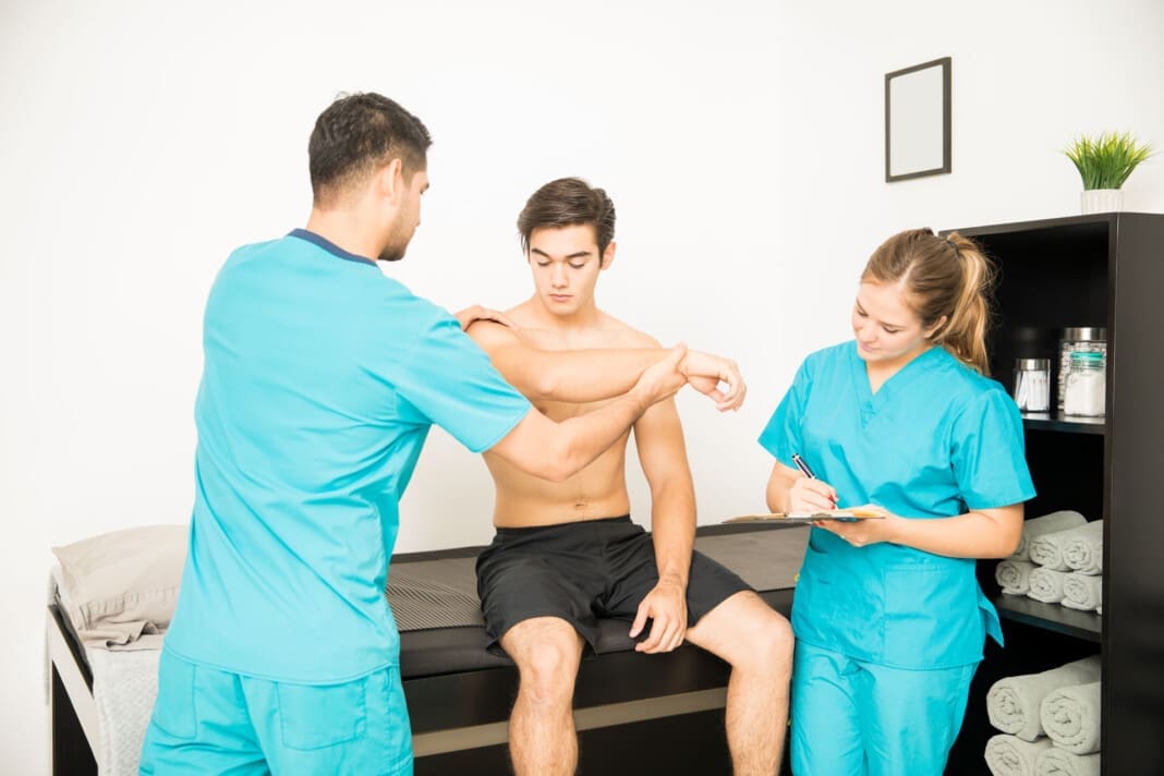 Physiotherapist Examining Shirtless Customer's Hand While Coworker Writing Notes