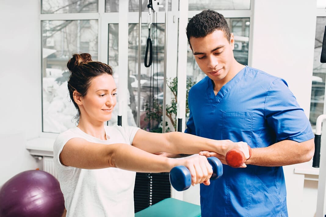 woman correctly performs exercise in the gym for the treatment of arthritis of the hands. The physiotherapist oversees the patient's exercise. Two dumbbells in the hands of a woman for exercise