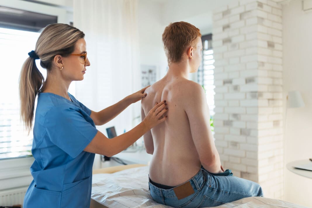An orthopedist examines the spine, posture, and spinal deformities of teenage patient. Teenage boy visiting paediatrician for annual preventive physical examination. Concept of preventive health care for adolescents.