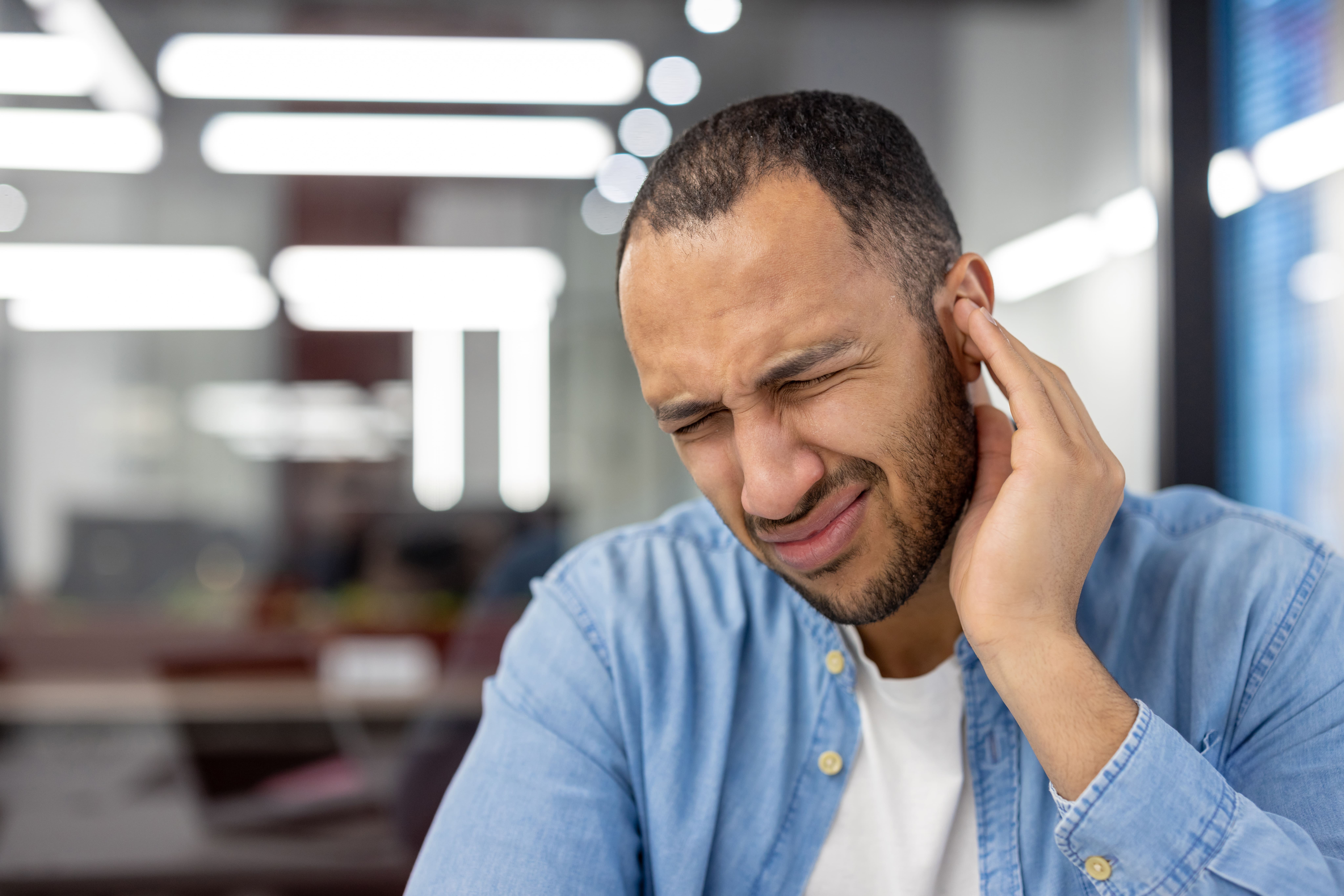 Close-up photo of a young Latin American man in the office, grimacing in pain, holding his ear with his hand